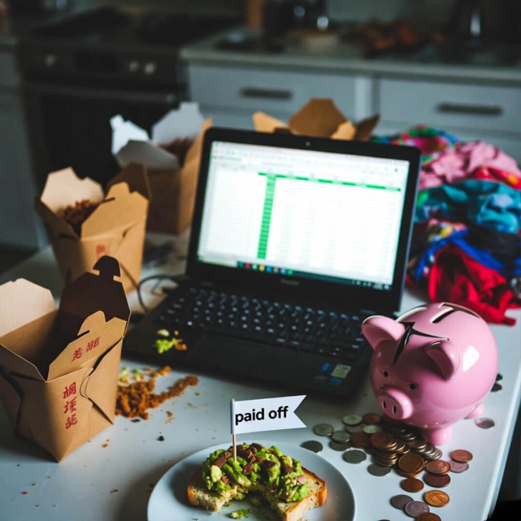 Cluttered table with cracked piggy bank, avocado toast, zero debt laptop.